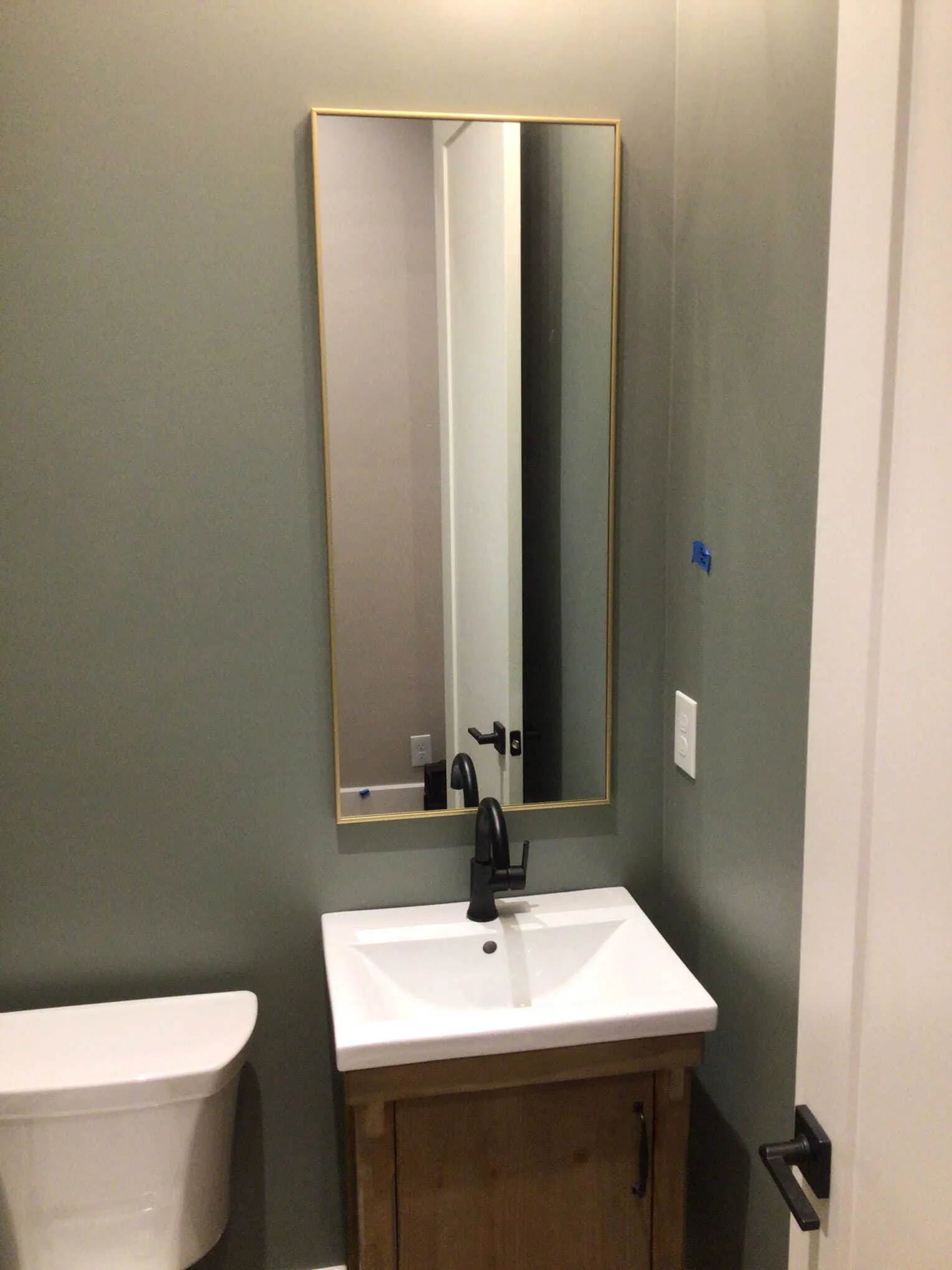 A small bathroom featuring a wooden vanity, a white sink with a black faucet, and a rectangular mirror with a gold frame.