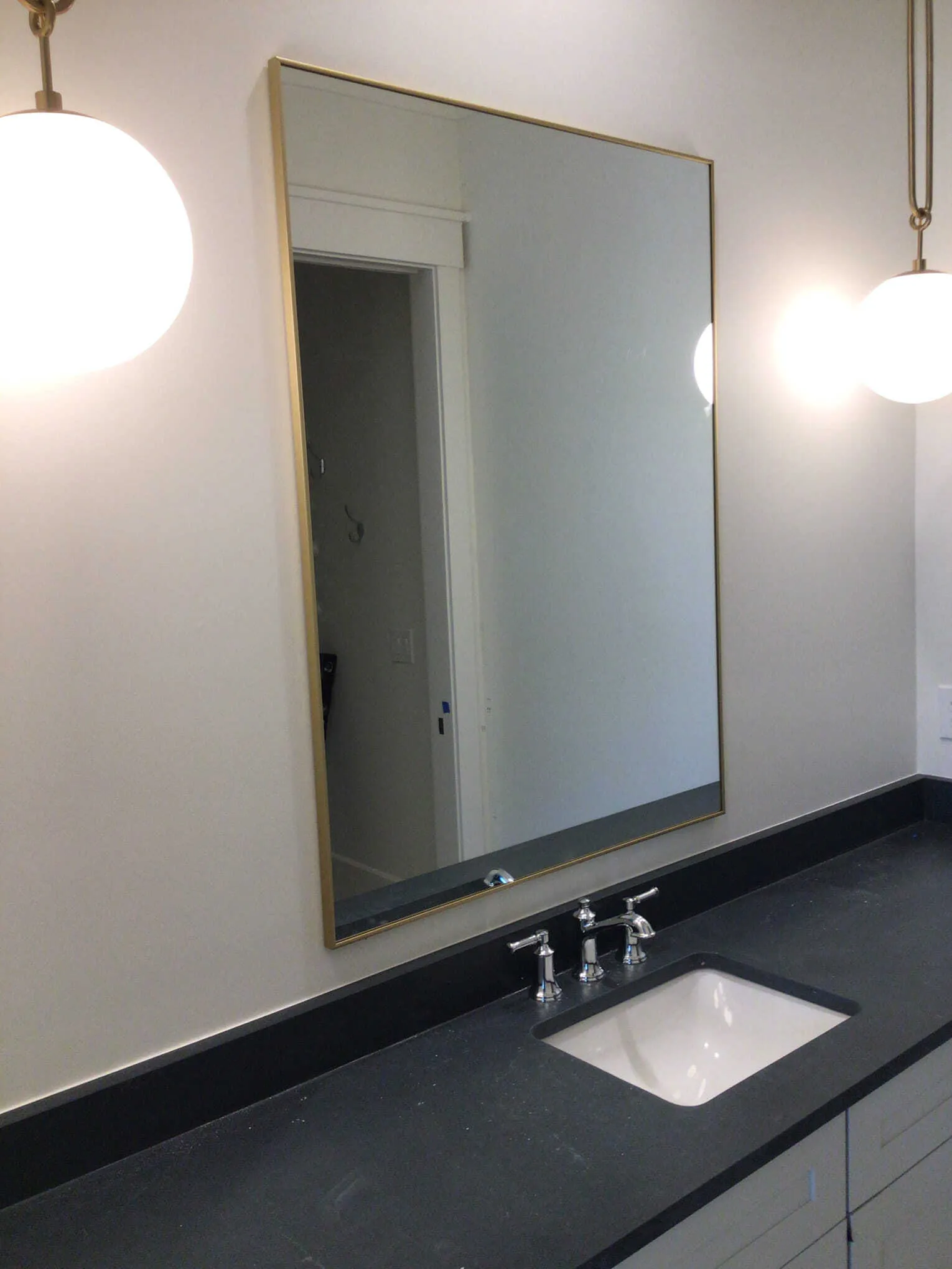 A bathroom vanity with a white sink, black countertop, rectangular gold-framed mirror, and two hanging globe lights.