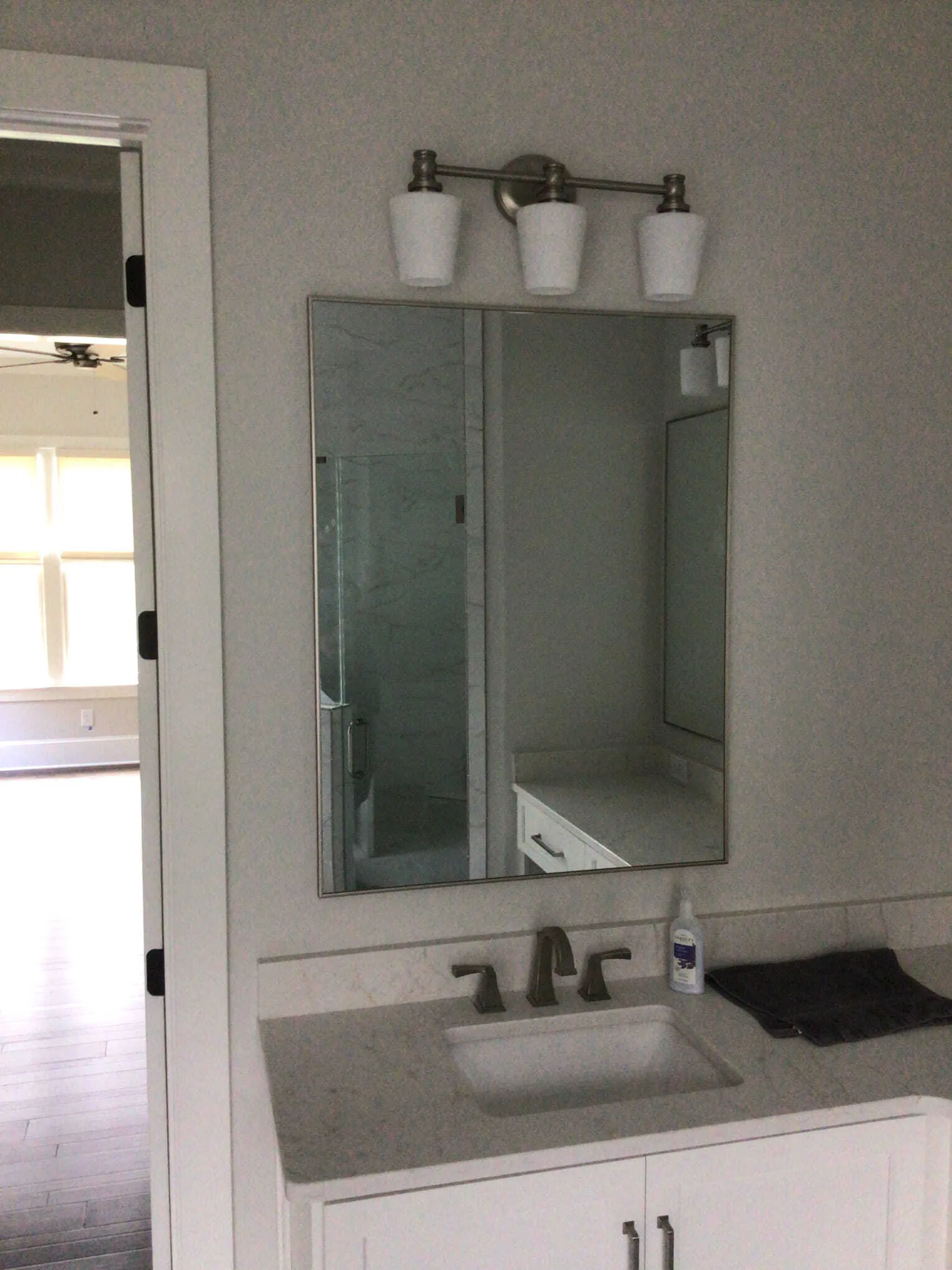 A white bathroom vanity with a rectangular mirror, three-light fixture, and open doorway in a neutrally-colored room.