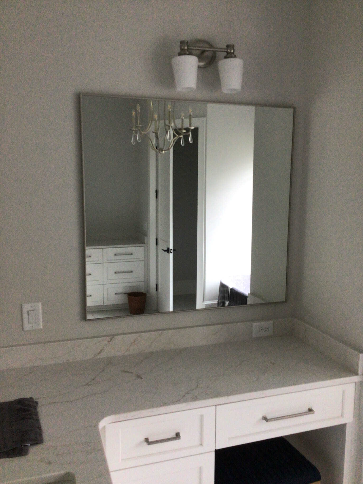 A wall-mounted square mirror hangs above a white bathroom vanity with marble-patterned countertops and a light fixture.