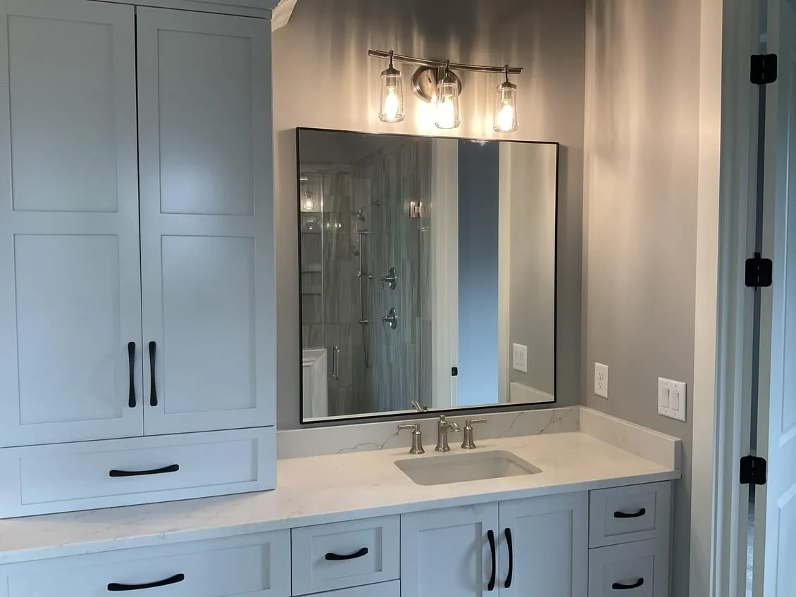 A bathroom vanity with white cabinets, dark hardware, a quartz countertop, a rectangular mirror, and a three-light fixture.