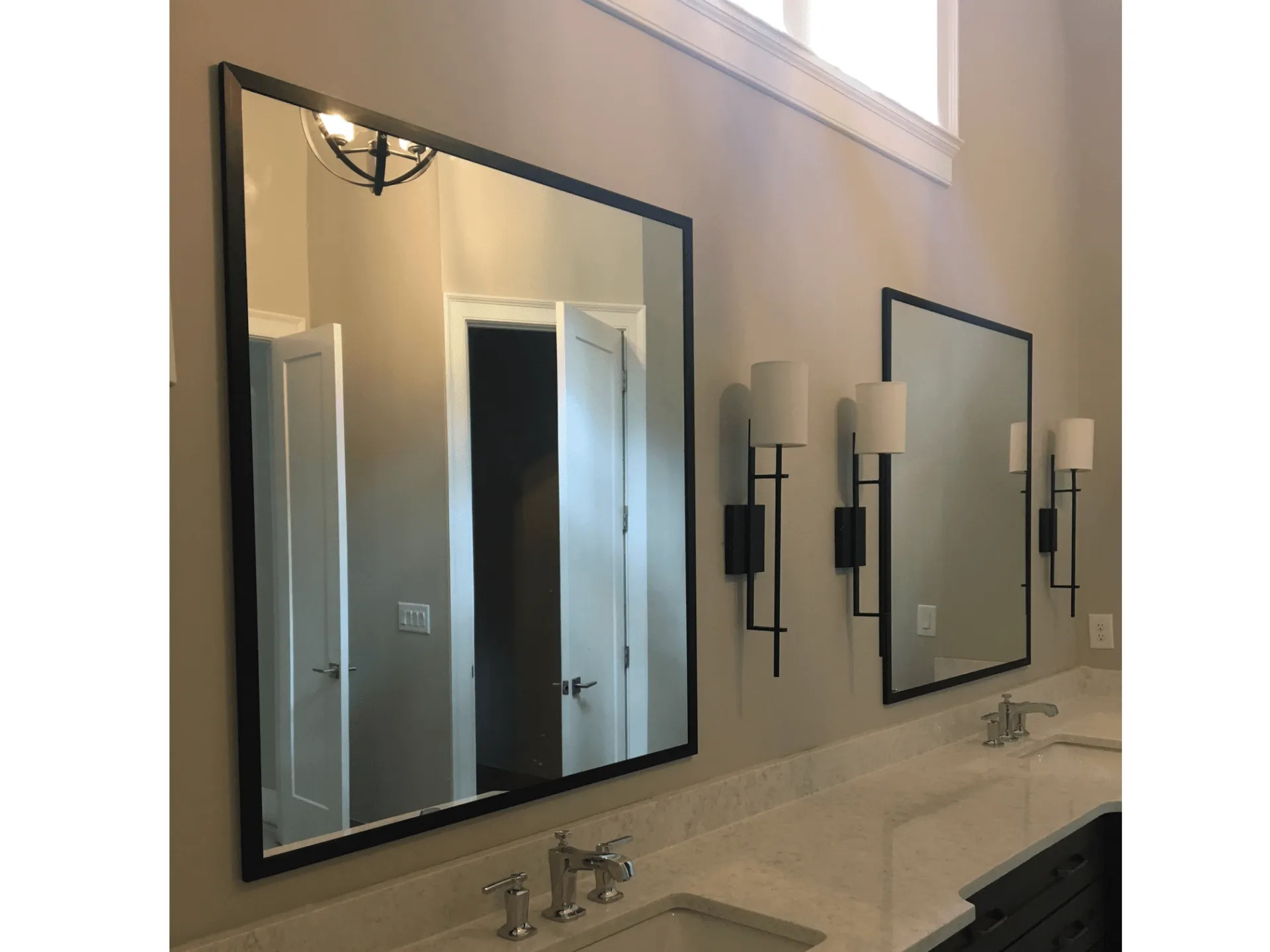 Two rectangular bathroom mirrors with black frames mounted on a beige wall above a white vanity with two sink faucets.