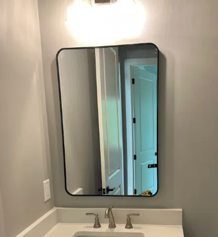 Black-framed rectangular mirror over a bathroom sink. The mirror reflects a doorway, sink, and light fixture. Gray walls.