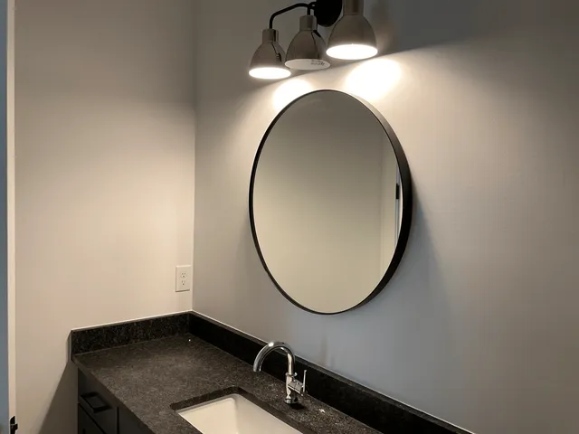 A bathroom vanity featuring a black-framed round mirror, dark stone countertop, white undermount sink, and three-bulb lamp.