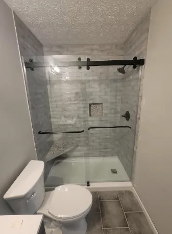 Bathroom with a glass shower enclosure, gray tiled walls, and a toilet. Black hardware.