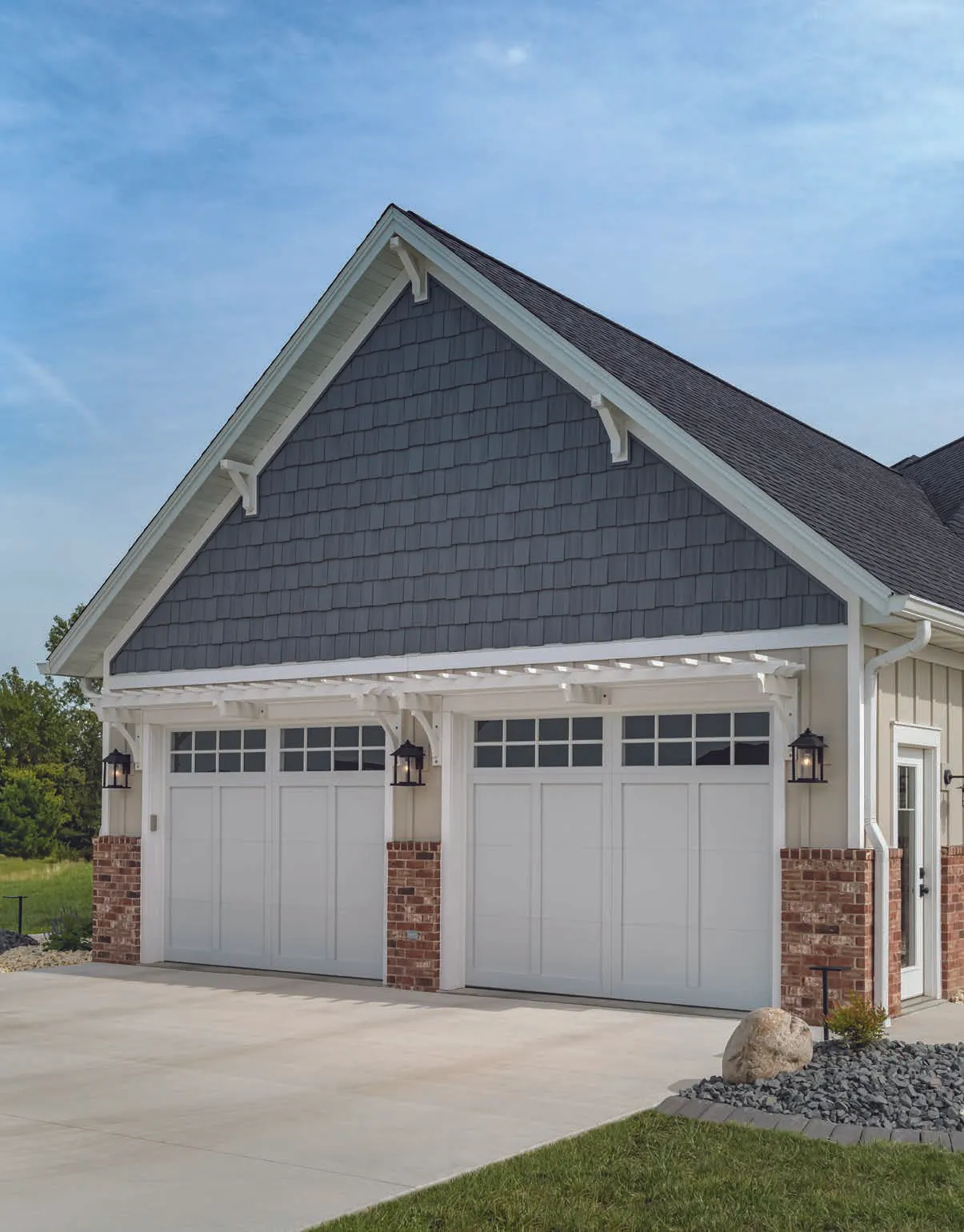 A detached two-car garage with white doors, dark gray shingle siding, and brick accents under a blue…