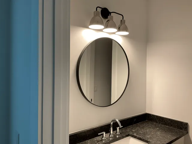 A circular mirror with a black frame hangs above a dark granite bathroom vanity with a three-light fixture above it.