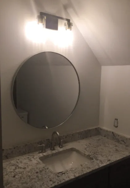 A round bathroom mirror mounted above a granite vanity with a sink and faucet, illuminated by a light fixture above.