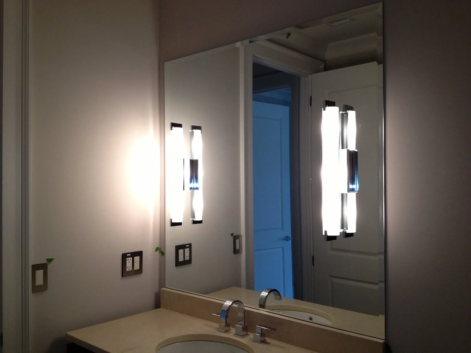 A bathroom vanity with a large wall mirror, two illuminated vertical light fixtures, and two electrical wall switches.