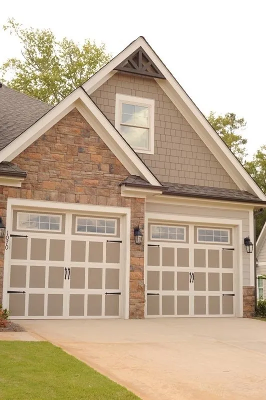Two-car garage with light-colored doors featuring windows, set in a stone and shingle home exterior under a peaked roof.
