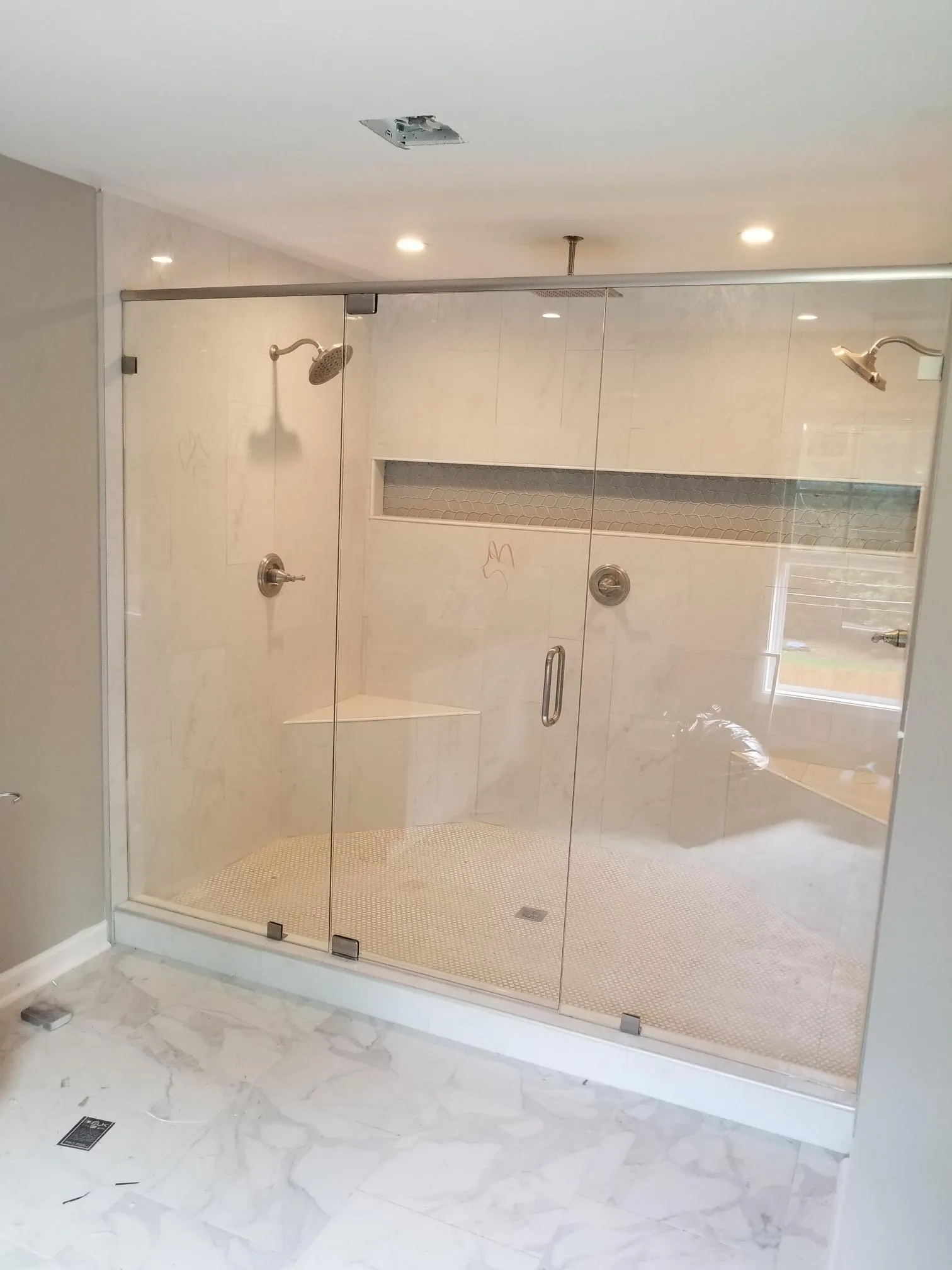 Glass shower enclosure with two showerheads, a seat, and stone tile walls.