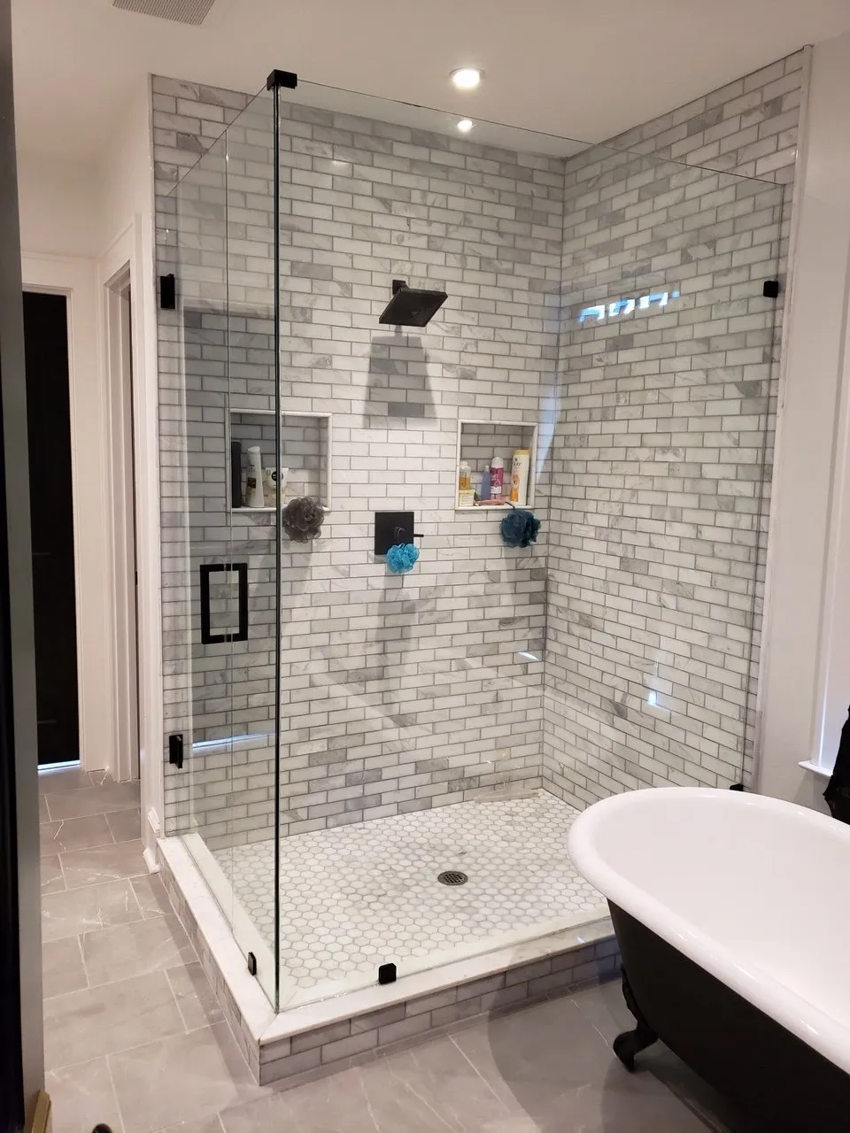 Glass shower with marble-look tile, black hardware, and pebbled floor. Window with blinds and patterned wallpaper visible.