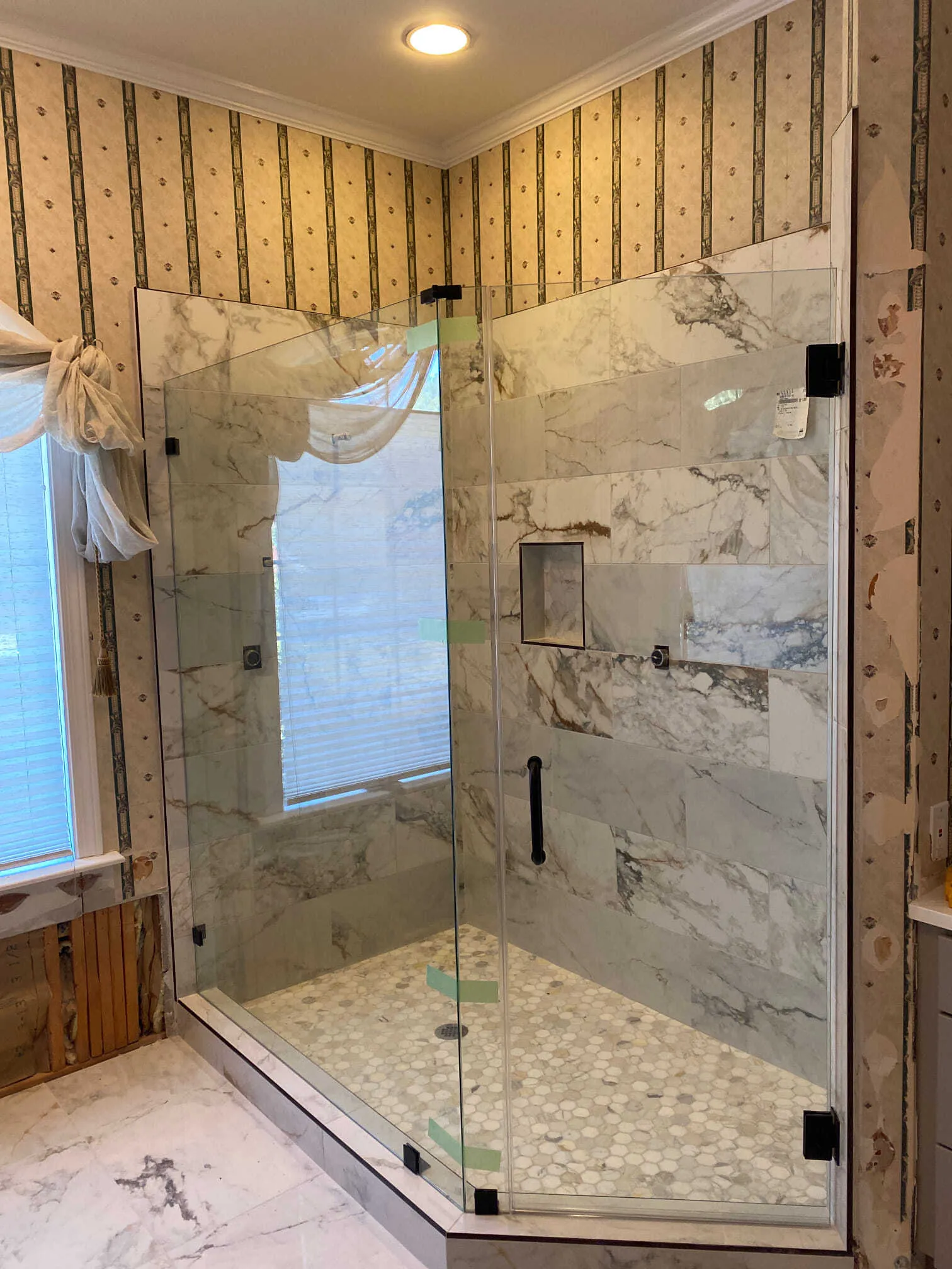 Glass shower with marble-look tile, black hardware, and pebbled floor. Window with blinds and patterned wallpaper visible.