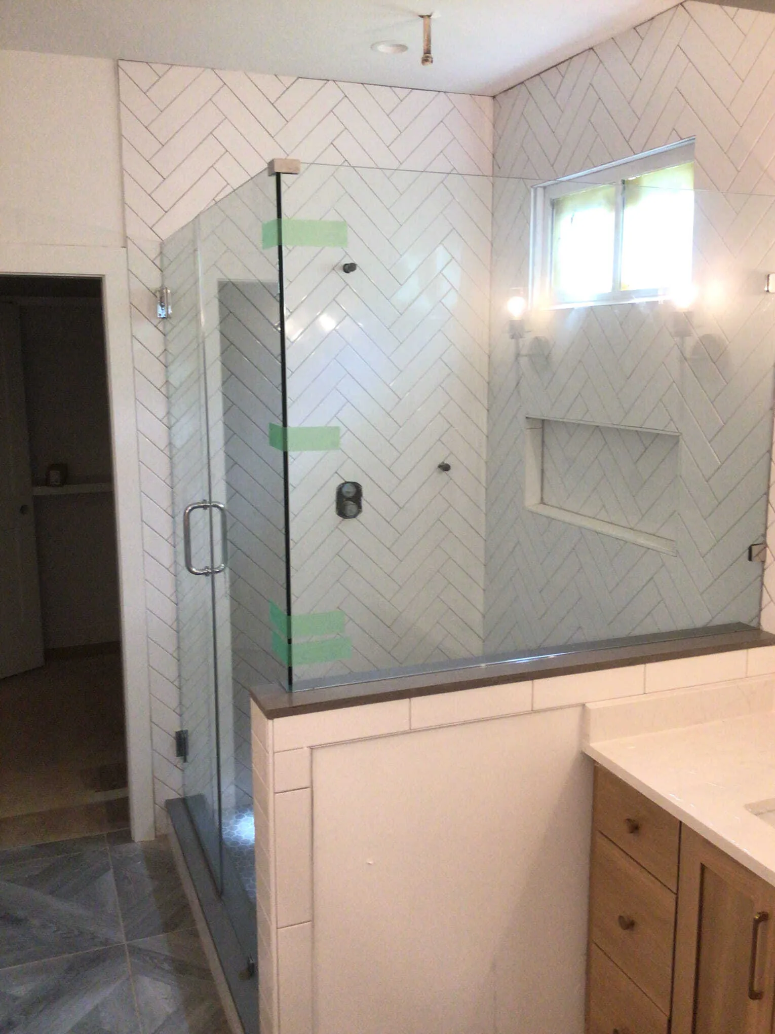 Corner glass shower with marble tile, gray cabinets, and a rug.