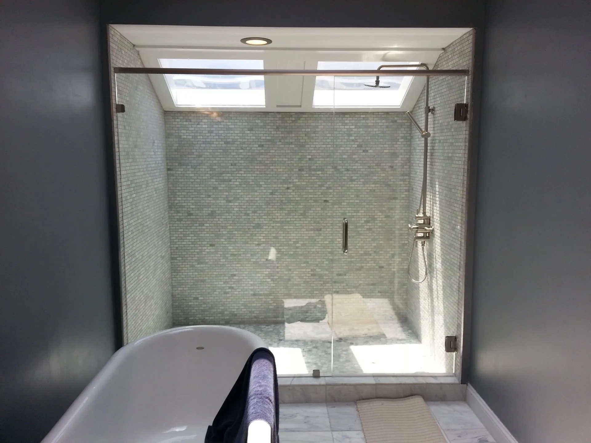 Shower with glass doors and mosaic tile, beside a bathtub in a room with dark gray walls and a skylight.