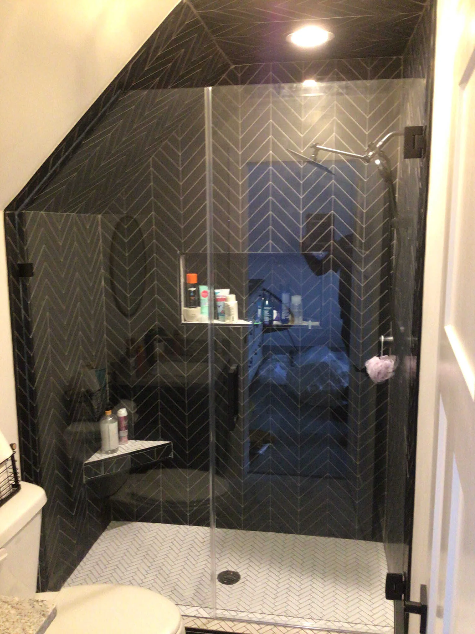 Modern shower with dark chevron-patterned tiles, glass door, white floor tiles, and a built-in shelf.