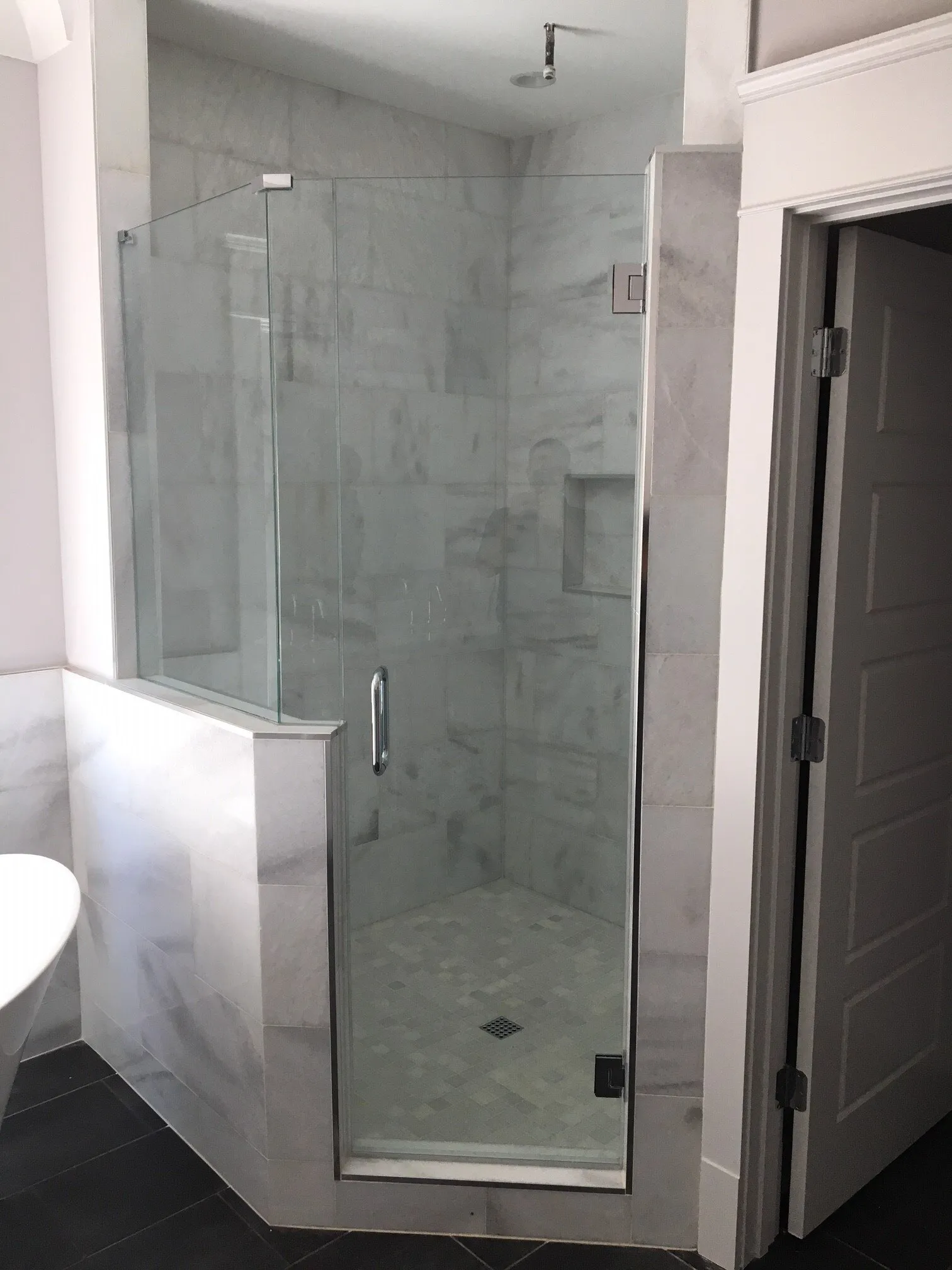 Glass-enclosed shower with white marble tile walls and dark floor. A door and an open doorway are visible.