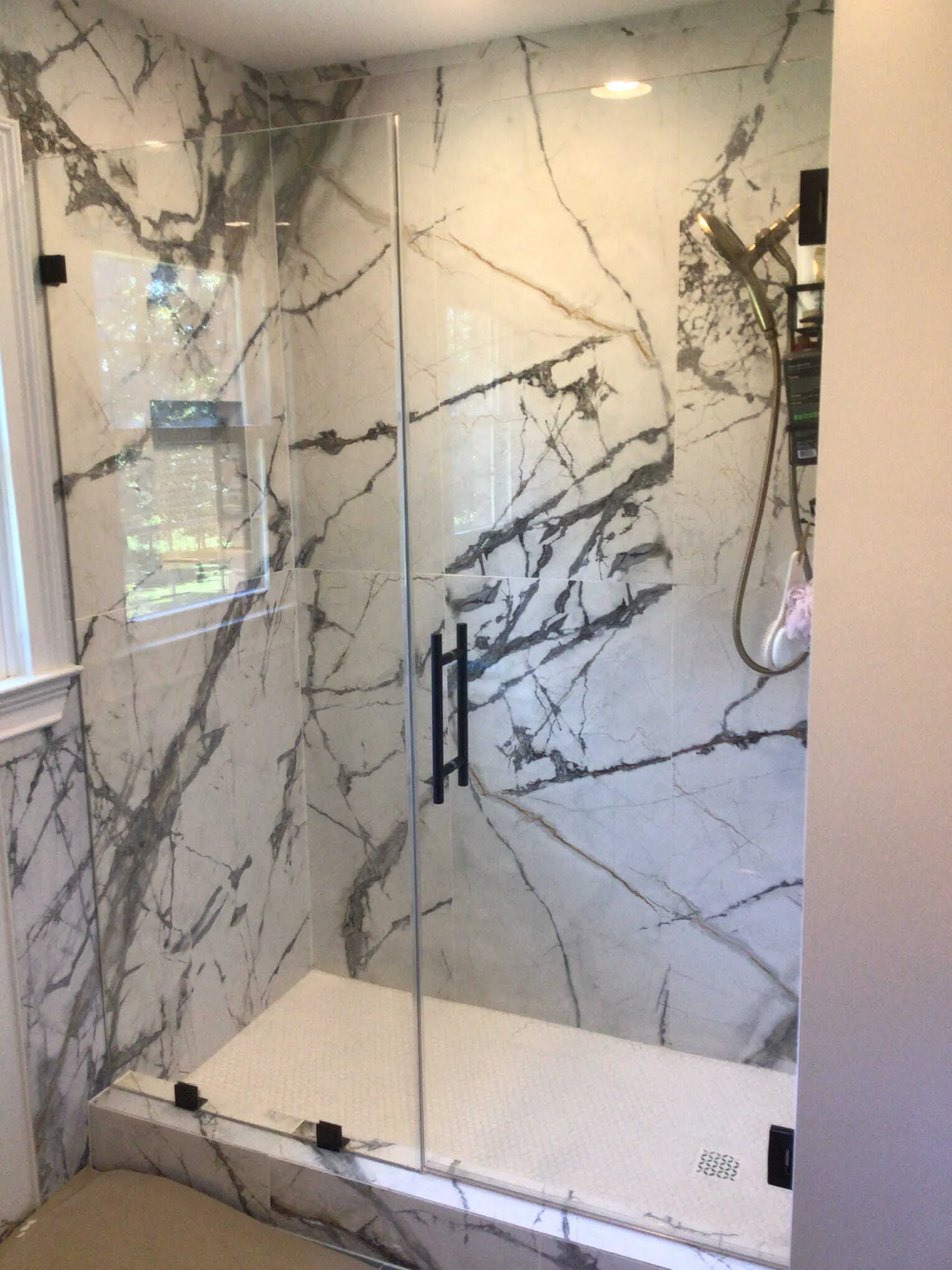 A marble-tiled shower with glass doors, black hardware, and a small window.