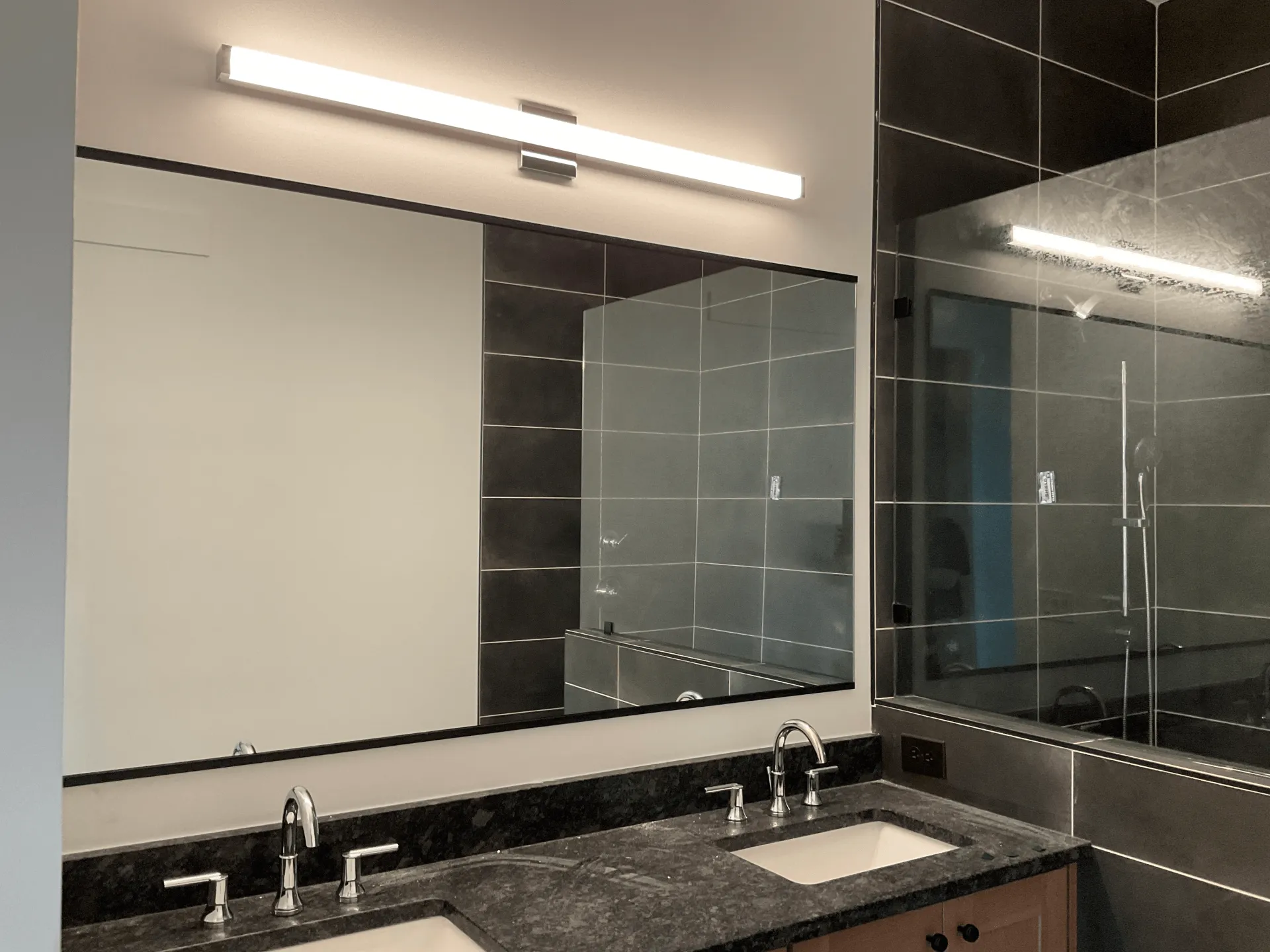 Bathroom with large mirror, double sinks, dark granite countertop, and linear light fixture.