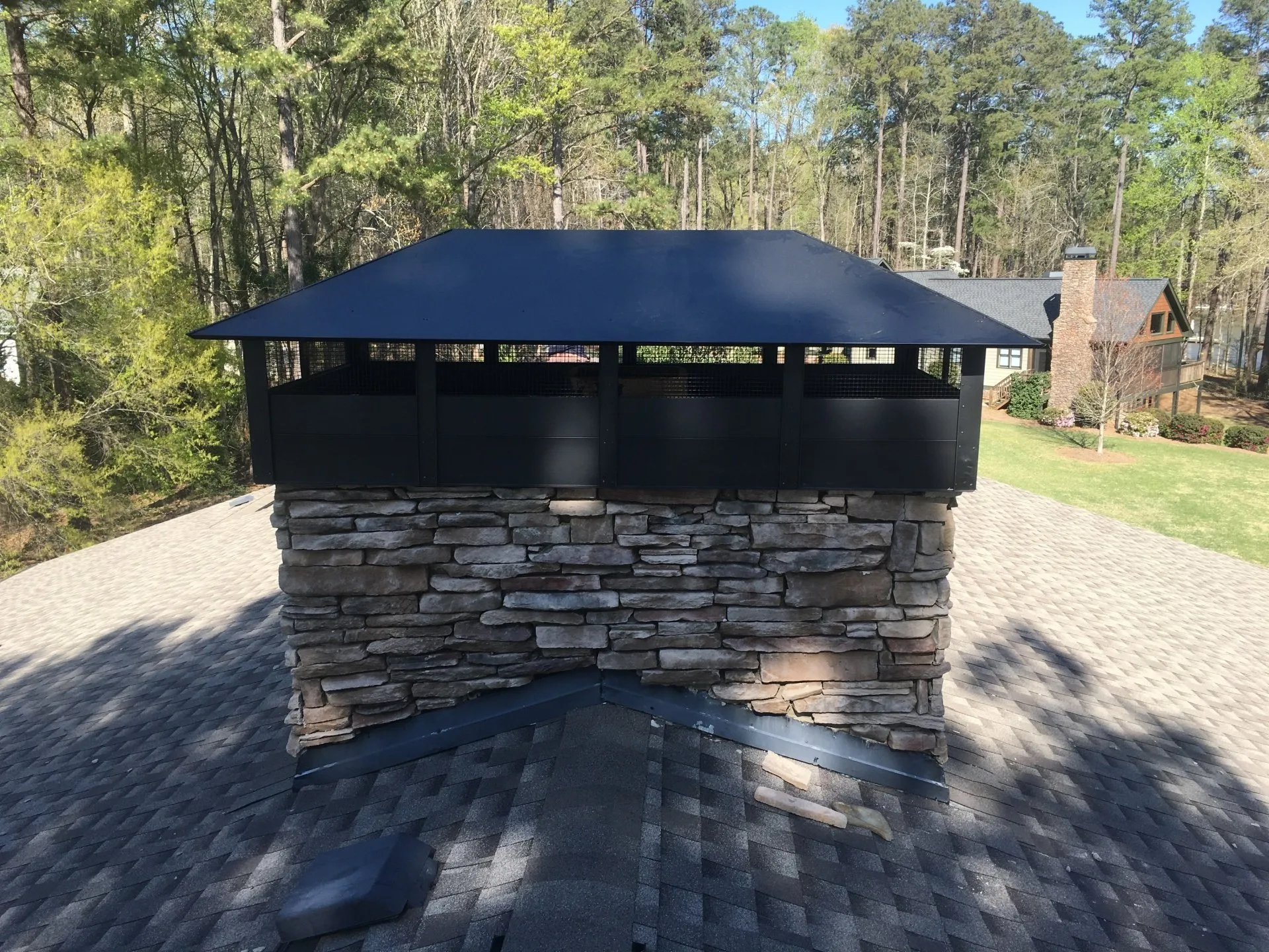 A stone chimney on a shingled roof features a large, black metal chimney cap against a background of trees.