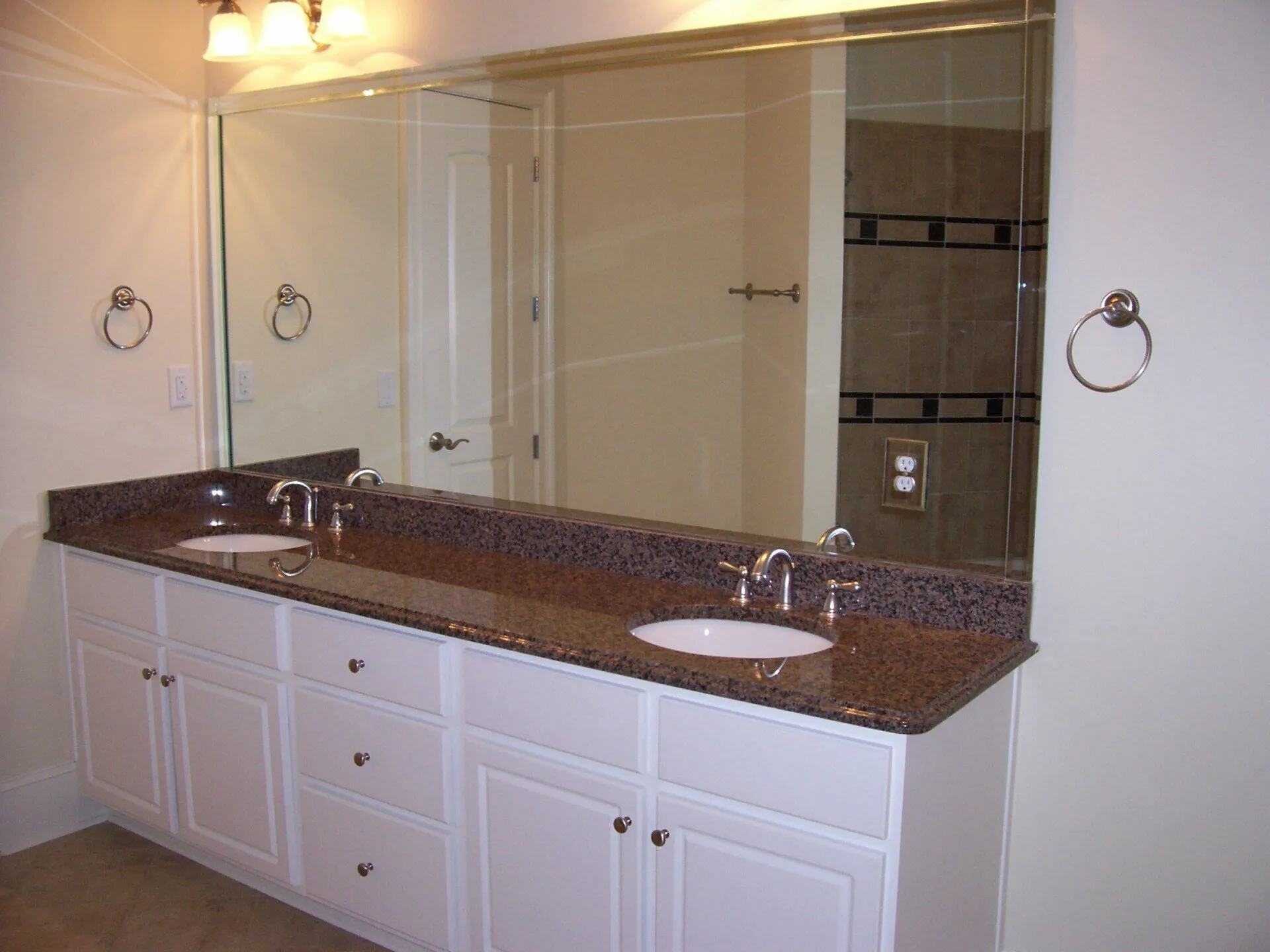 Double sink bathroom vanity with white cabinets, speckled brown granite countertop, and a large wall-to-wall mirror.