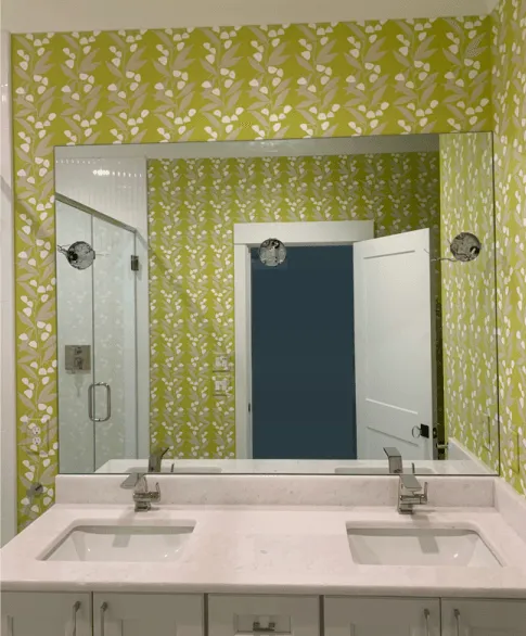 A double-sink bathroom vanity with light countertops and white cabinets against a lime green floral wallpaper.