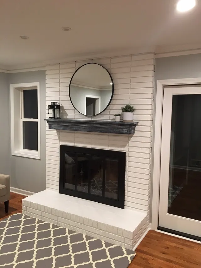 White brick fireplace with a dark wooden mantel, circular mirror, and black glass doors in a room with a patterned rug.