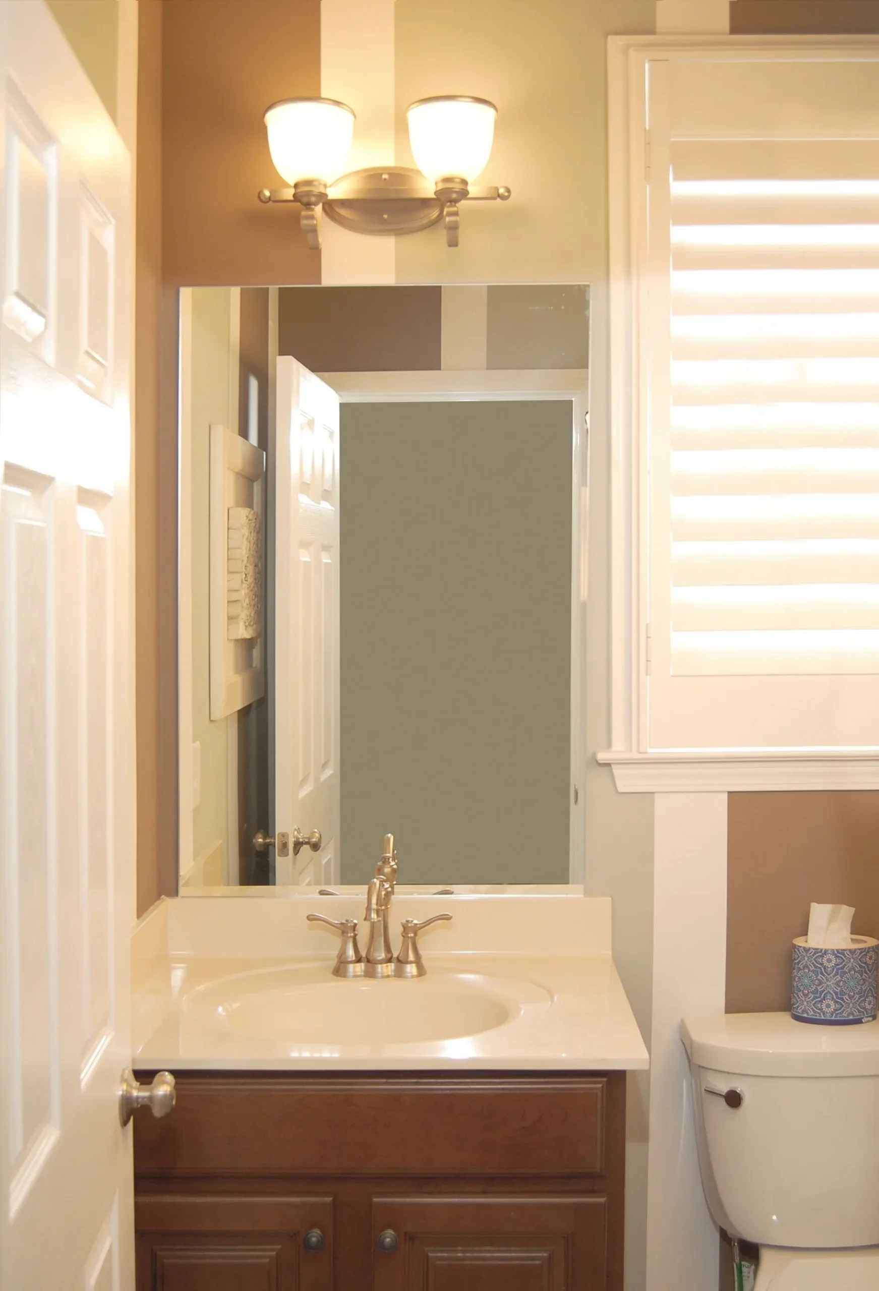 A bathroom vanity with a wooden cabinet, white sink, and silver faucet beneath a mirror, wall light, and window shutters.