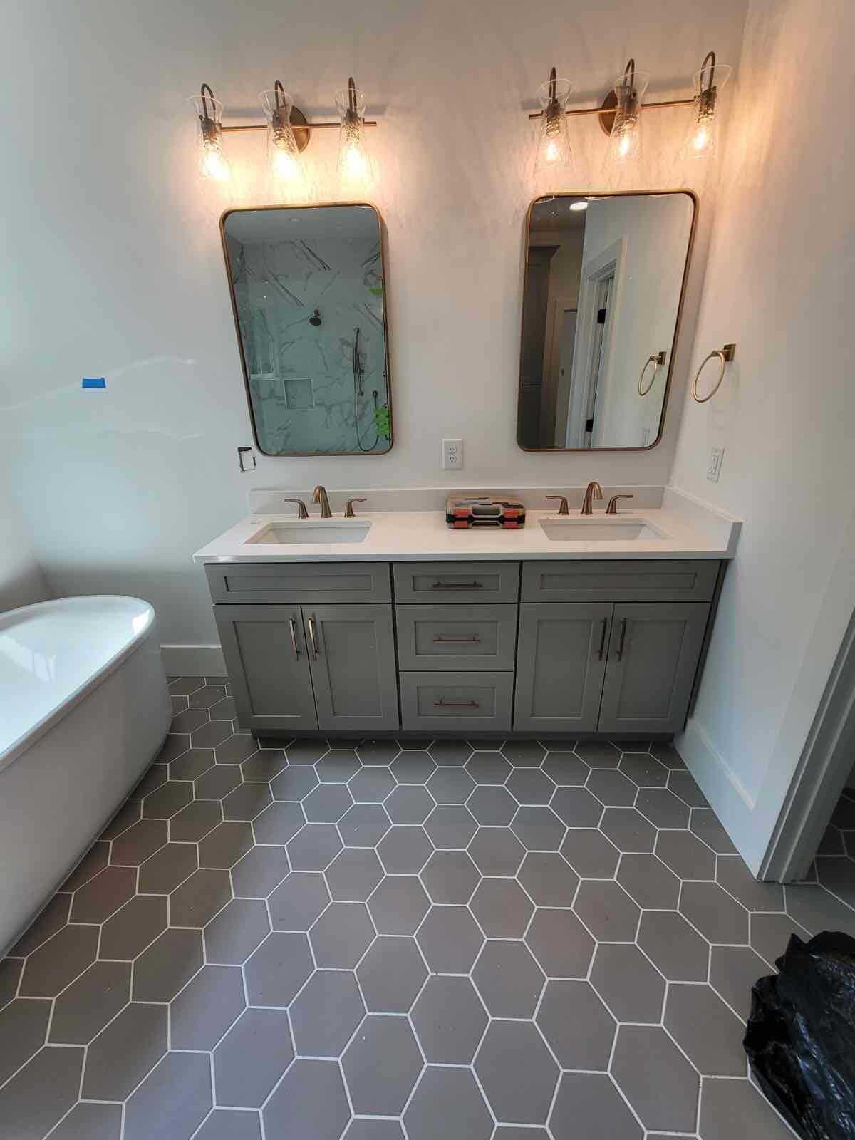 Bathroom with grey vanity, two sinks, mirrors, hexagon tile floor, and a bathtub.