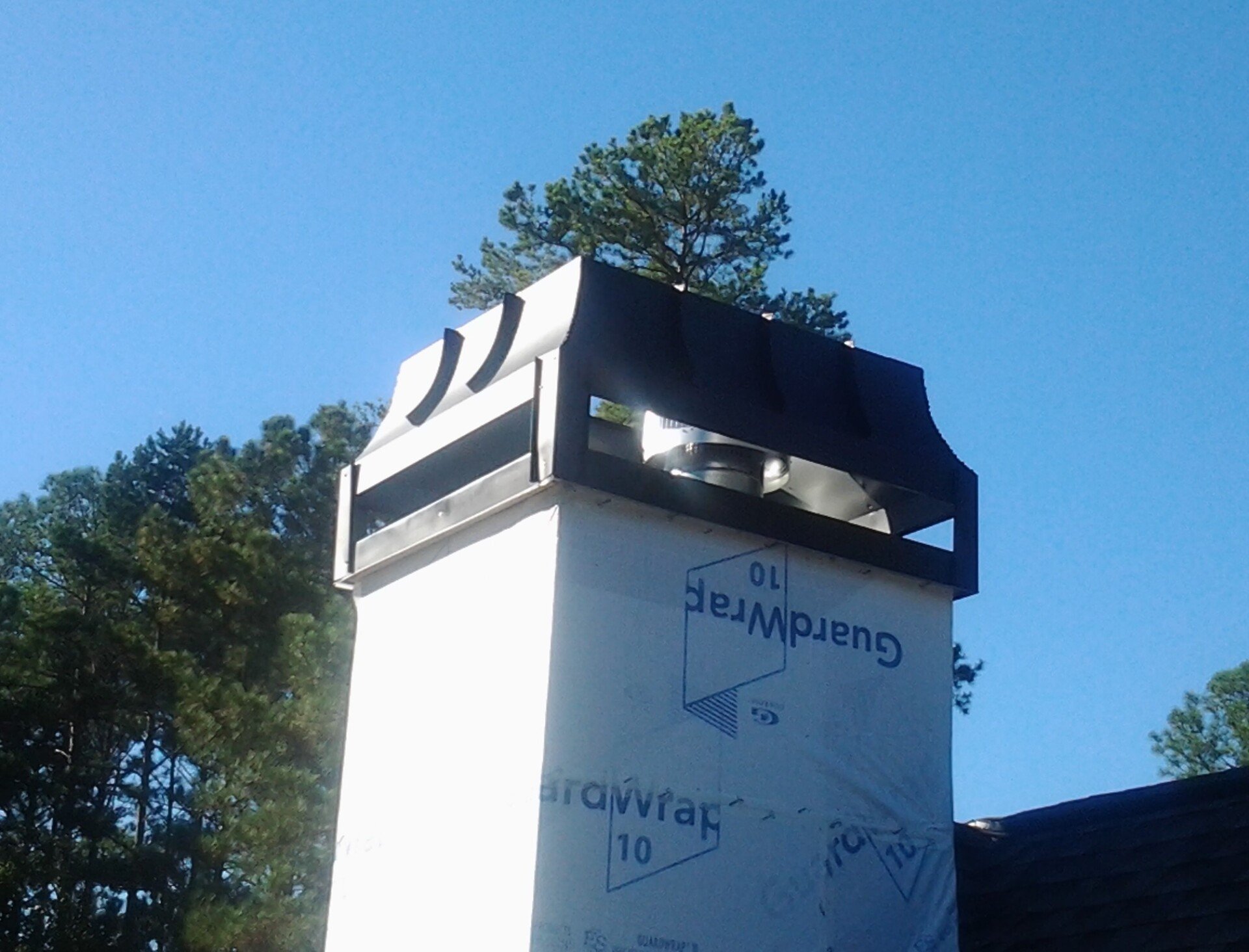 A chimney top under construction, wrapped in white GuardWrap building material against a bright blue sky.