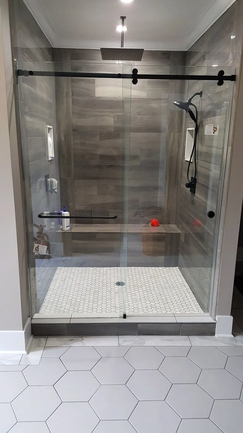 Modern shower with glass door, stone floor, built-in bench, dark fixtures, and wood-look tiled walls.