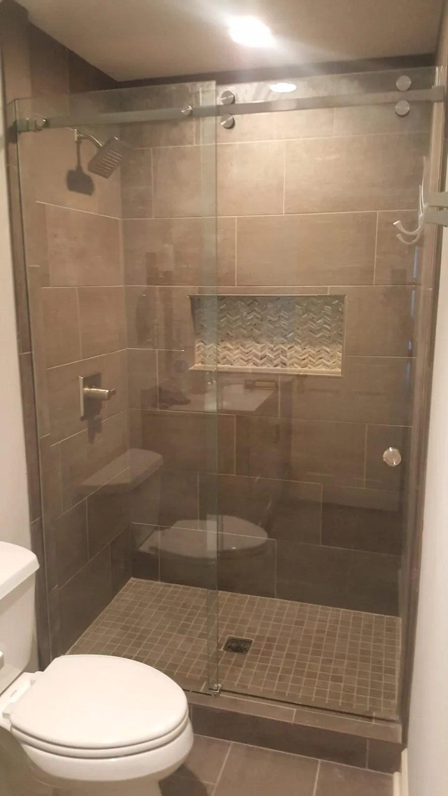 Shower with glass doors, brown tiled walls, built-in shelves, and a seat. Toilet is in the foreground.