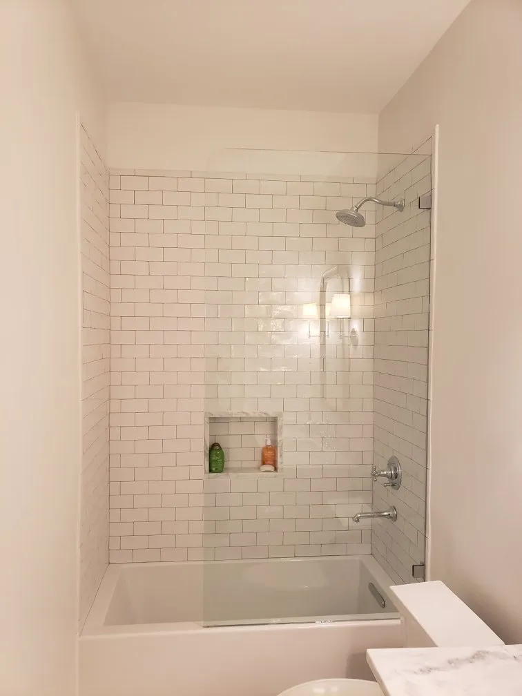 White-tiled shower with glass door and built-in shelf; chrome fixtures.