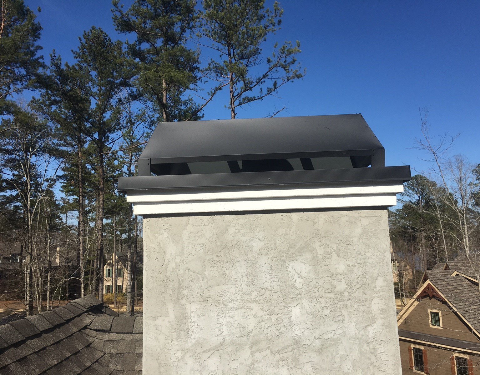 A gray stucco chimney top with a black metal shroud and white trim against a clear blue sky and pine trees.
