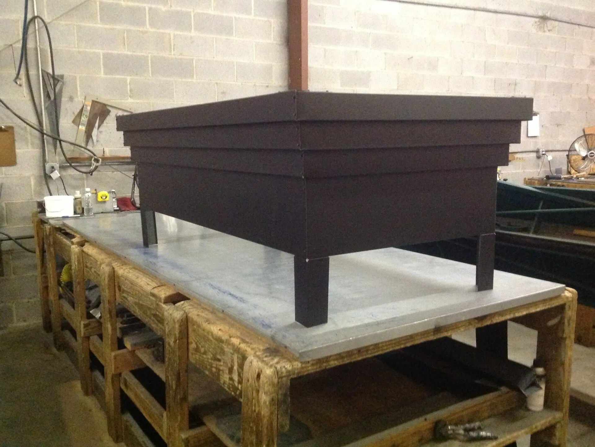A dark, rectangular tiered planter box sits on a wooden workbench inside a workshop with a cinder block wall background.
