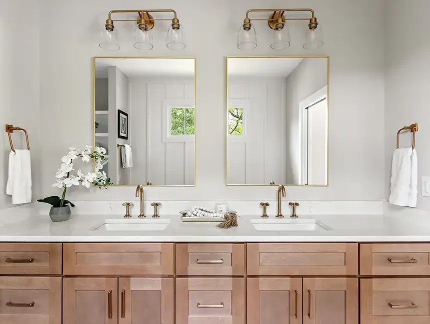 Modern bathroom vanity with light wood cabinets, white countertops, two sinks, mirrors, and brass fixtures.