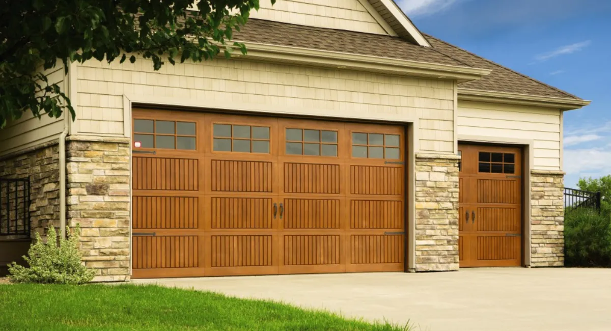 Brown garage doors with windows and stone accents on a beige house with green grass.