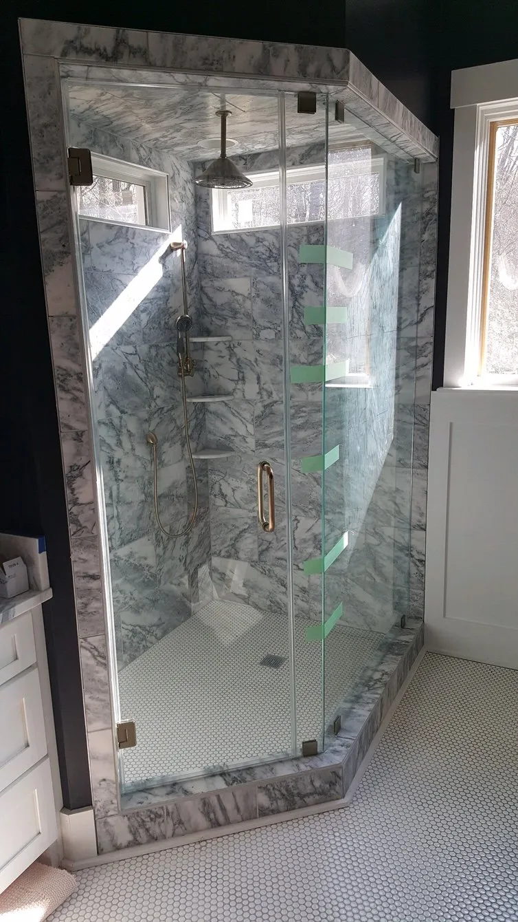Marble-tiled shower with glass doors and a square shower head, set on a mosaic-tiled floor.