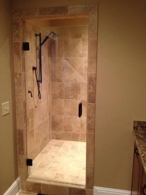Walk-in shower with tiled walls and floor, framed by stone. Glass door, black hardware, and bronze shower fixtures.