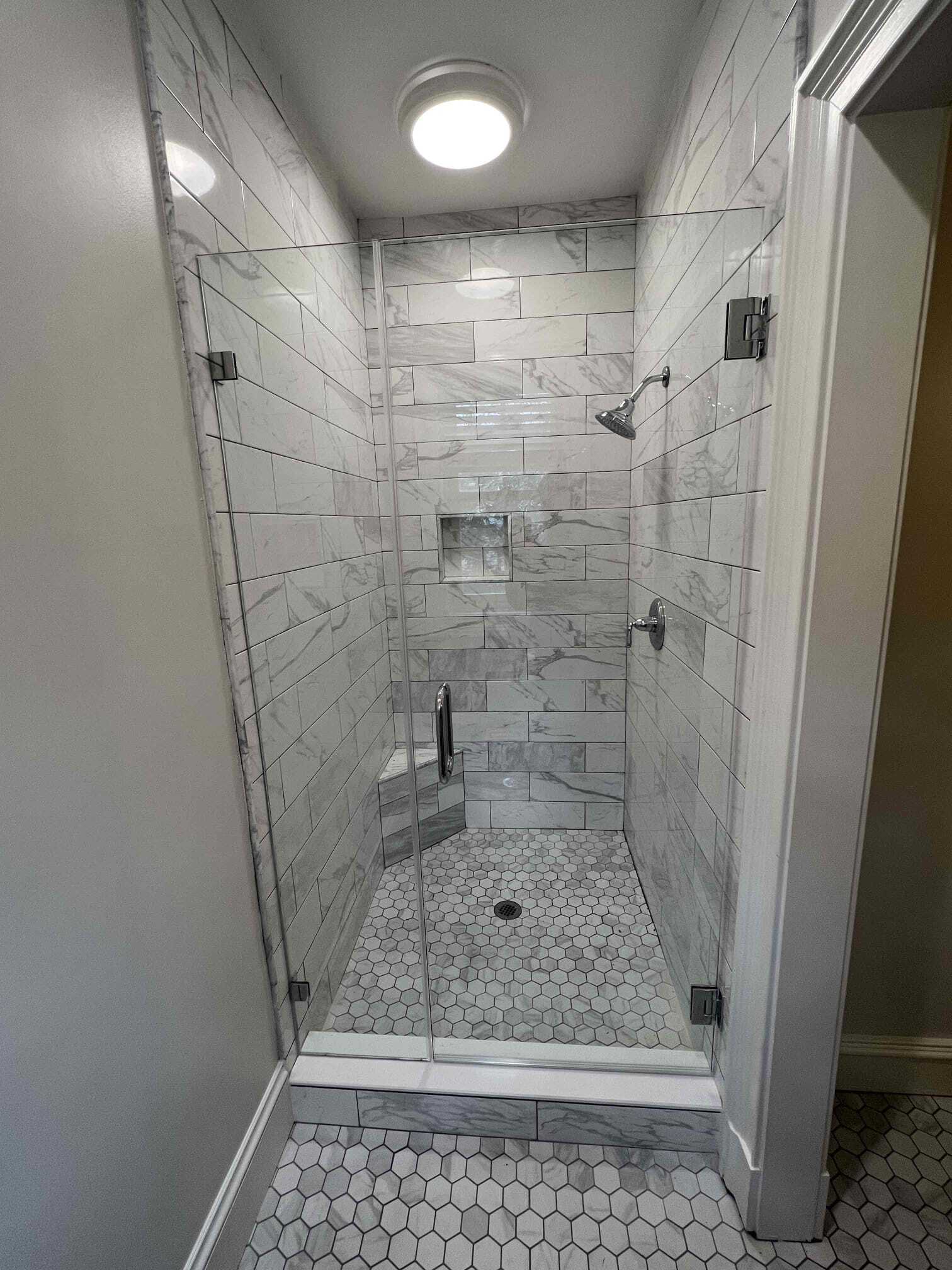 Glass-enclosed shower with white marble tile walls and dark floor. A door and an open doorway are visible.
