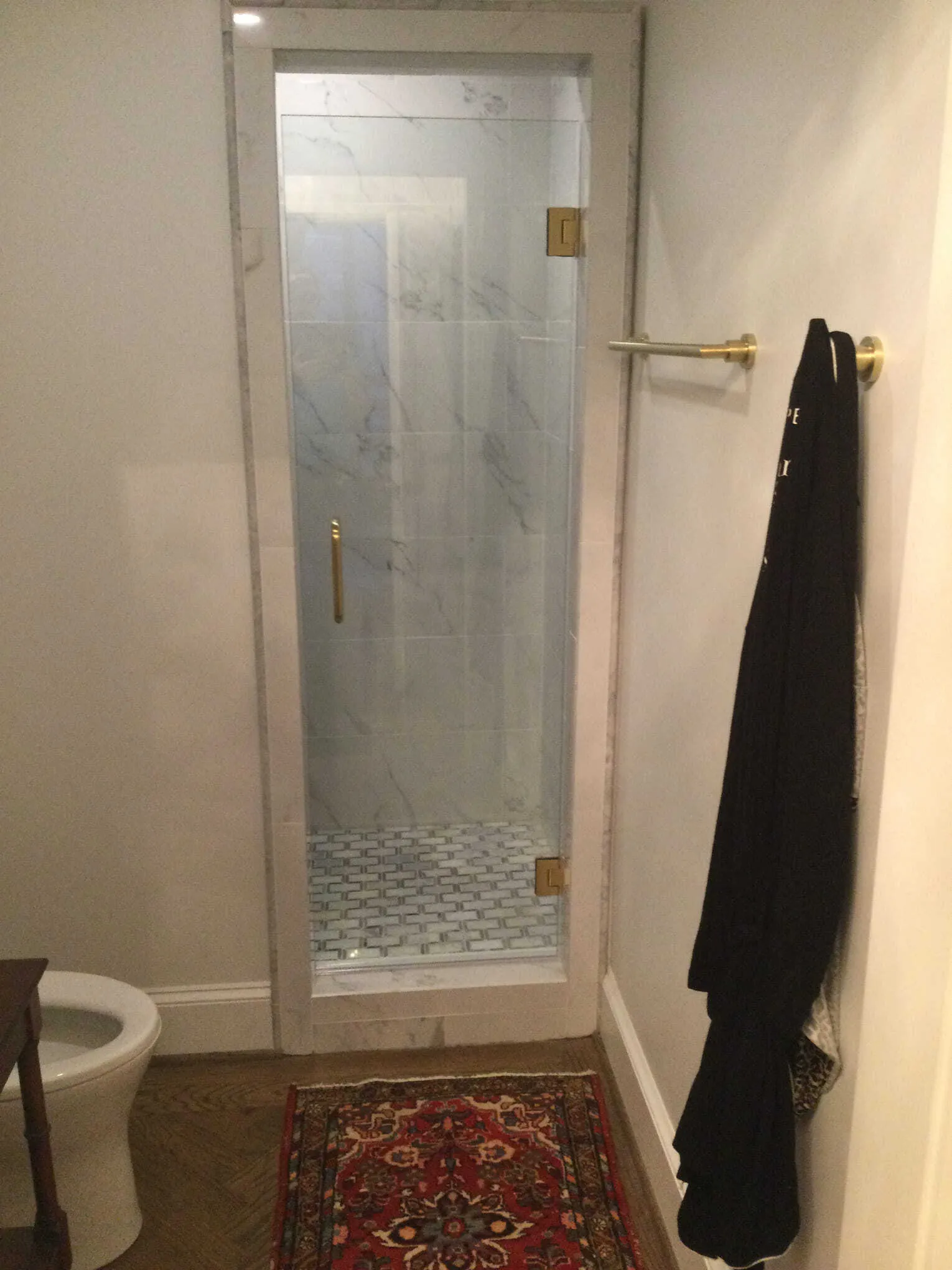 Bathroom with a glass shower door, gold fixtures, and a black robe hanging on a wall. A small rug is on the floor.