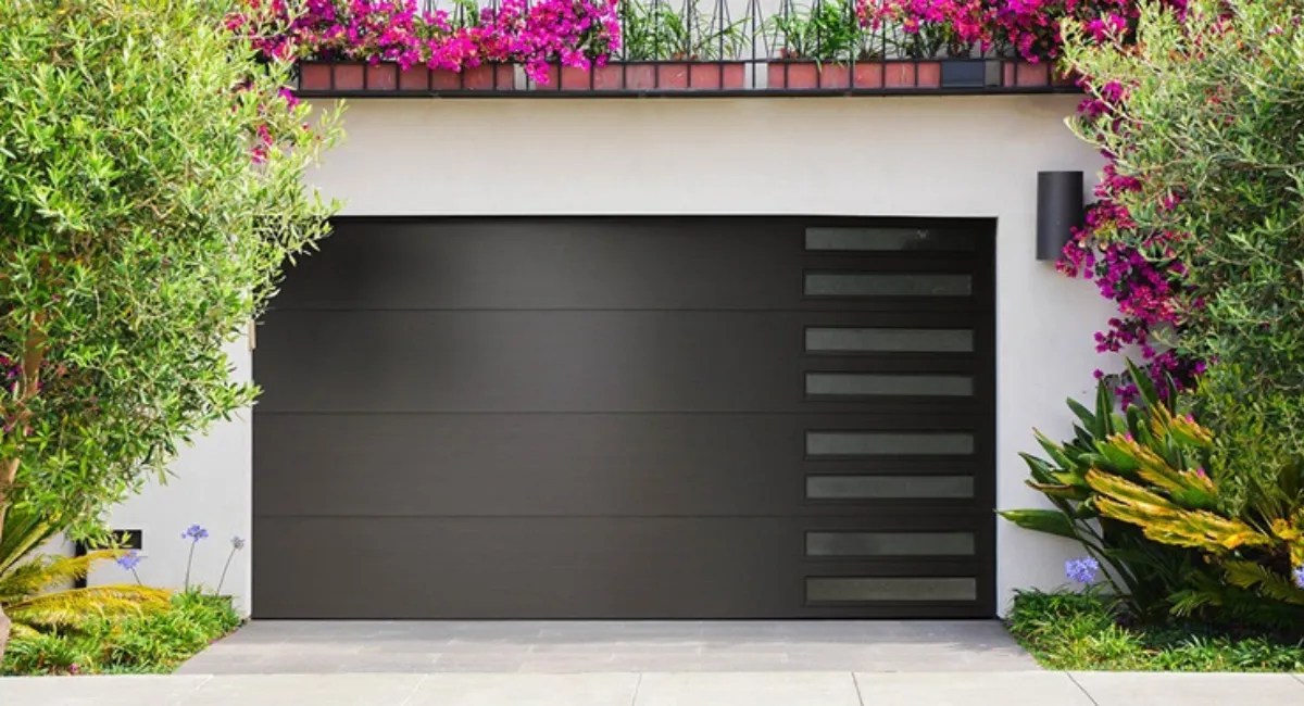 Modern black garage door with horizontal panels and rectangular windows, surrounded by green foliage and pink flowers.
