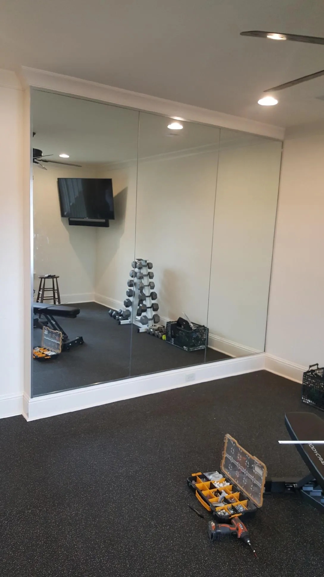Large wall mirrors installed in a home gym with dumbbells, a bench, and a toolbox on a dark rubber floor.