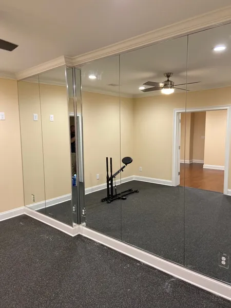 A home gym room with mirrored walls, a black exercise machine on speckled flooring, and an open doorway to another room.