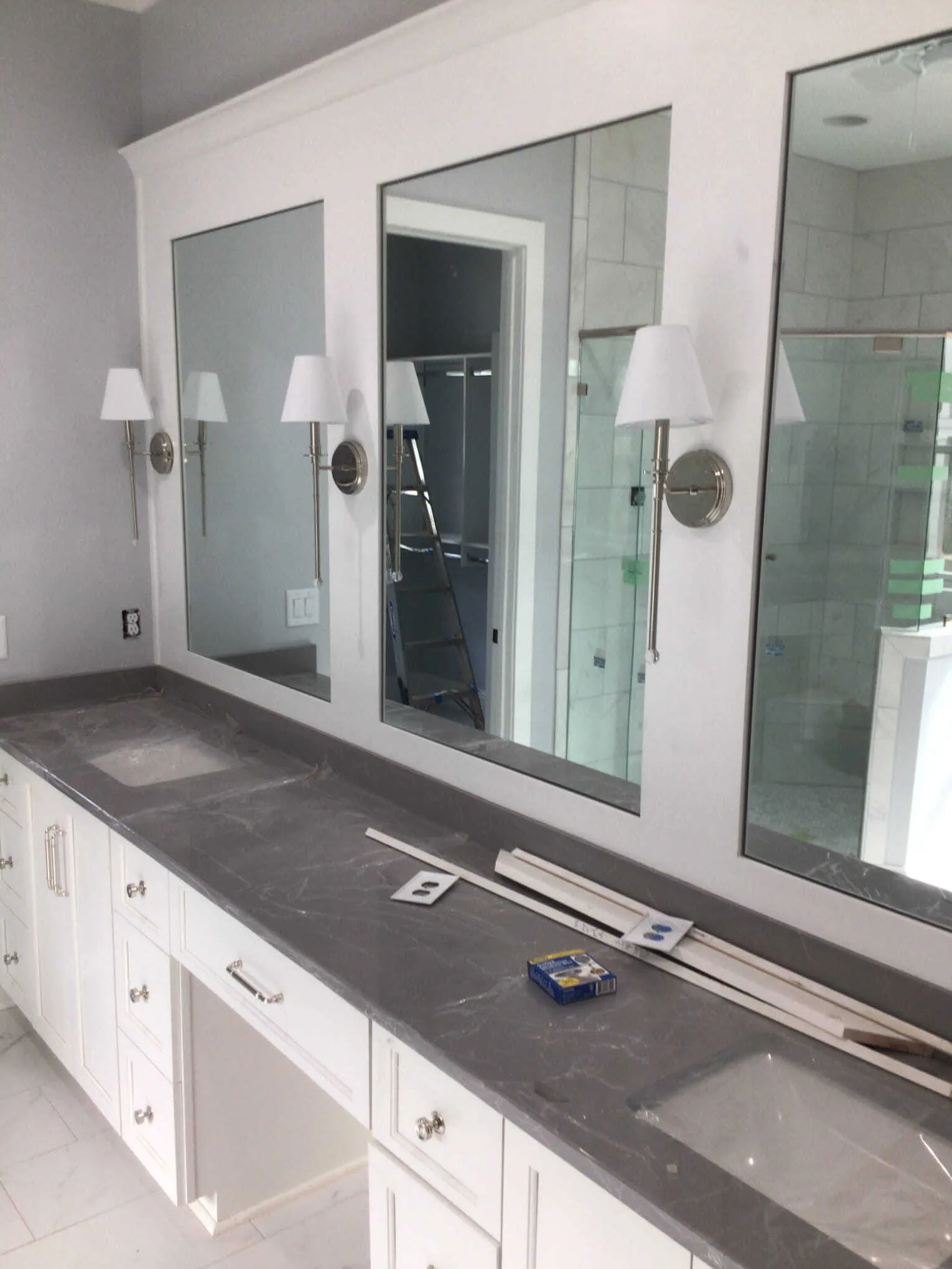 A white double-vanity bathroom with gray stone countertops, three framed mirrors, and wall-mounted sconces.