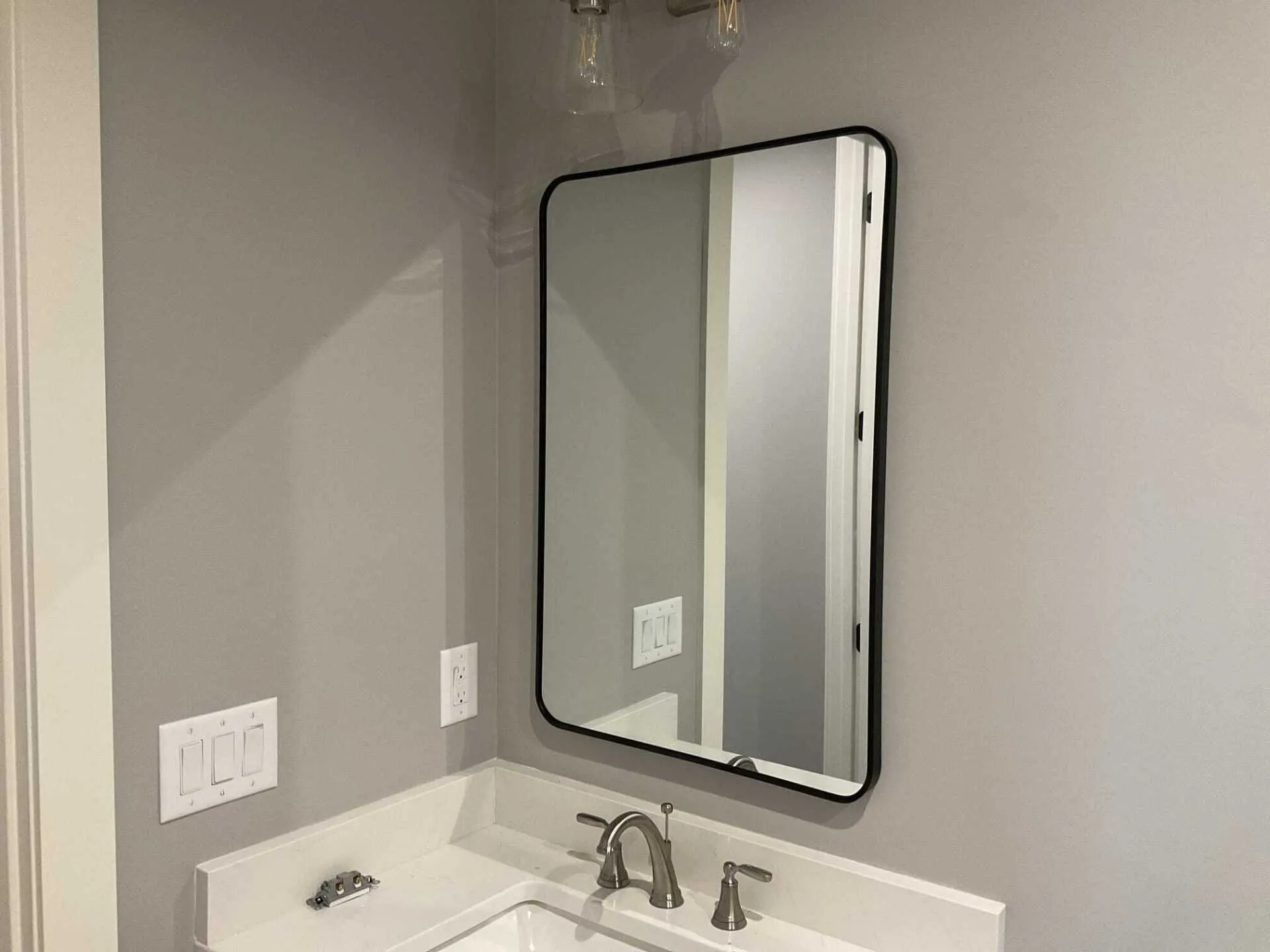 Black-framed rectangular mirror over a bathroom sink. The mirror reflects a doorway, sink, and light fixture. Gray walls.