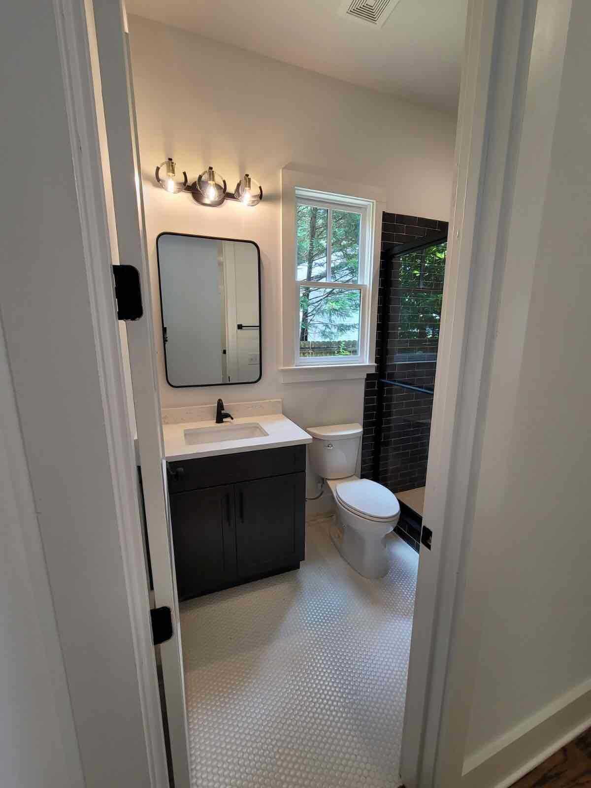 Bathroom with dark vanity, black-framed mirror, white toilet, and black-tiled shower, viewed from a doorway.