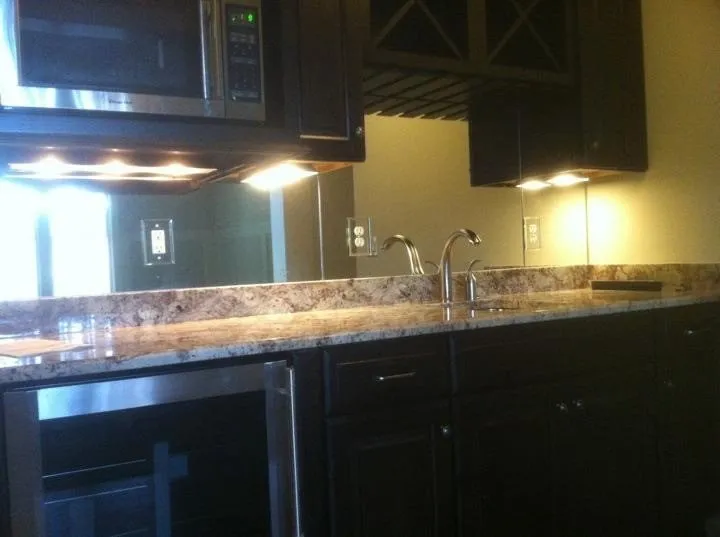 A wet bar with dark cabinetry, granite countertops, a built-in microwave, under-cabinet lighting, and a mirrored backsplash.